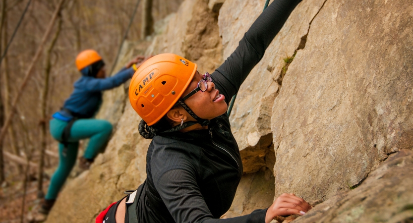 Two people wearing safety gear are secured by ropes as they each climb a rock face. 